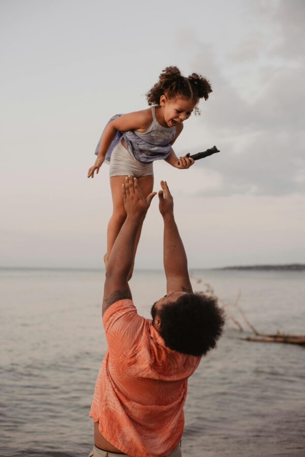 A joyful father lifts his daughter in the air by the beach during sunset, capturing a moment of love and happiness.